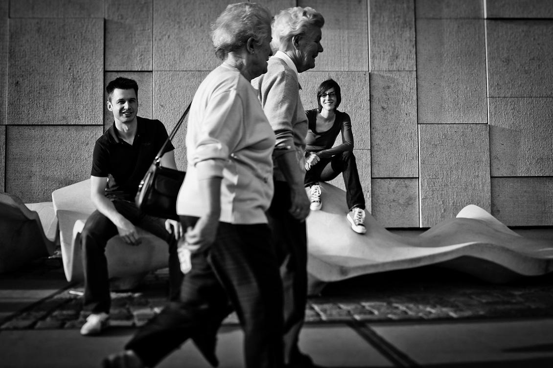 Engagement_shooting_hafencity_Hamburg_das_Paar_sitzt_auf_der_Skulptur_und_zwei_alten_Frauen_gehen_vorbei